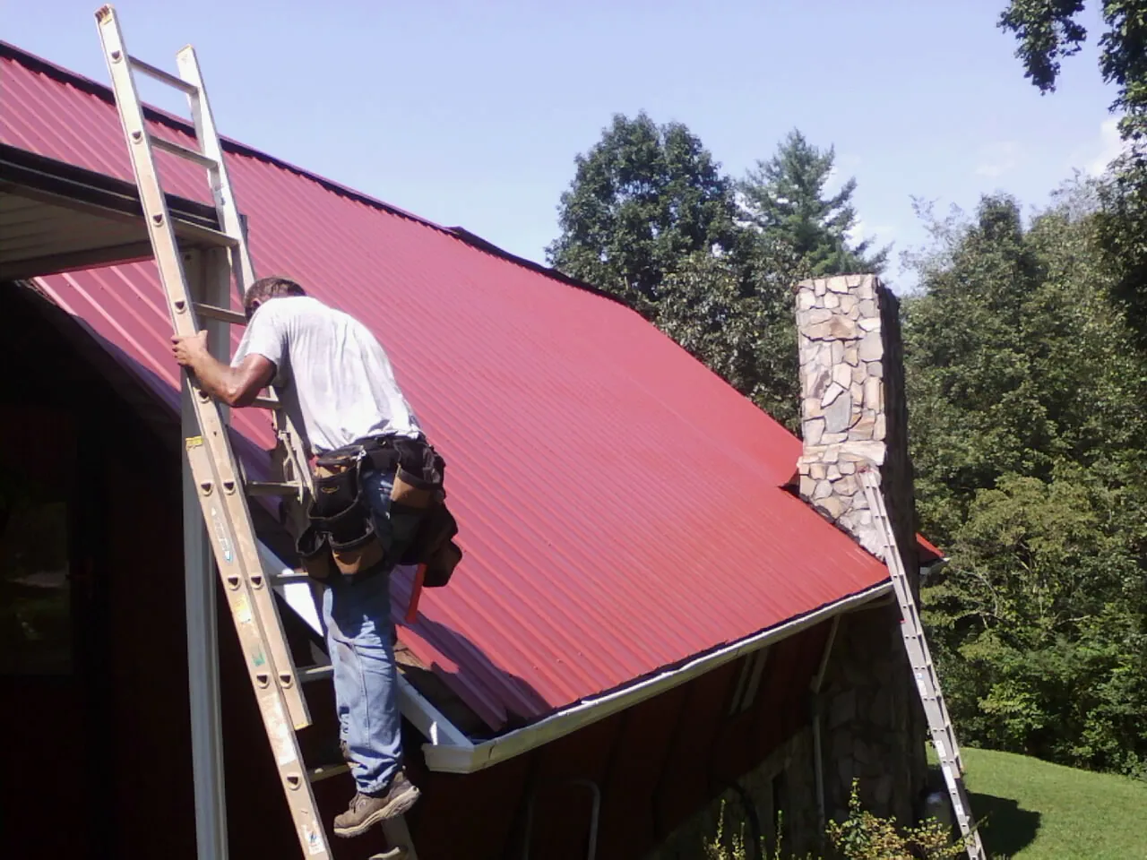 Roofer on a ladder beside a red metal roof and stone chimney — Soffit & Fascia Repair by Roofing Brothers in Hinsdale